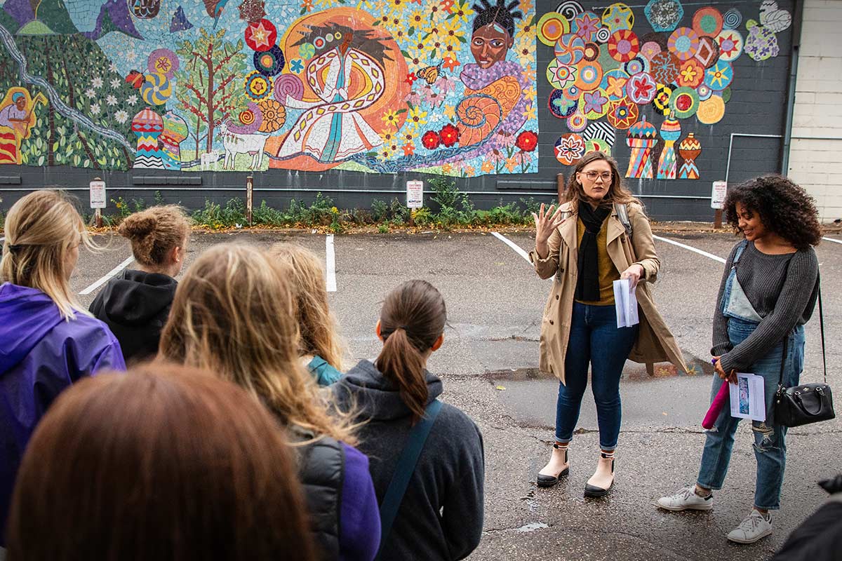 A teacher addressing a group of students in front of a mural.