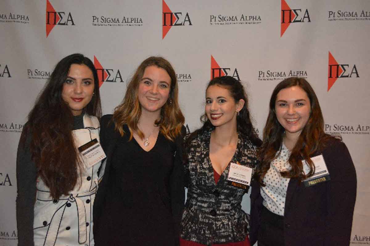 Four students pose for a photo in front a Pi Sigma Alpha Research Conference banner. 