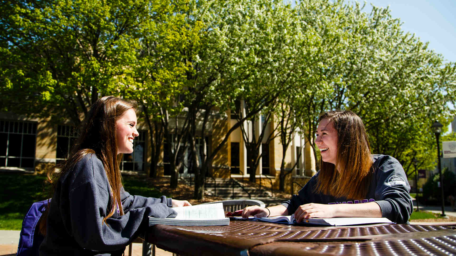 Two students sit and chat at a picnic table on a sunny day.