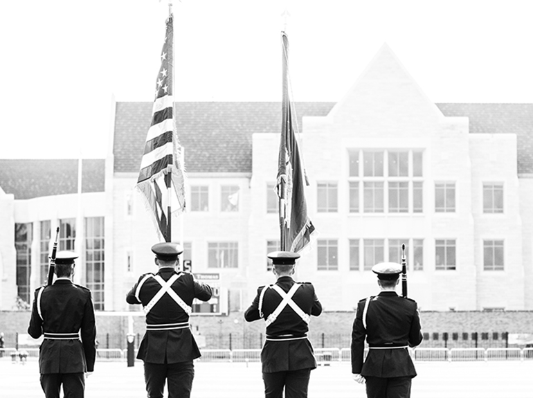 Four cadets march while performing an honor guard detail on the Saint Thomas football field.