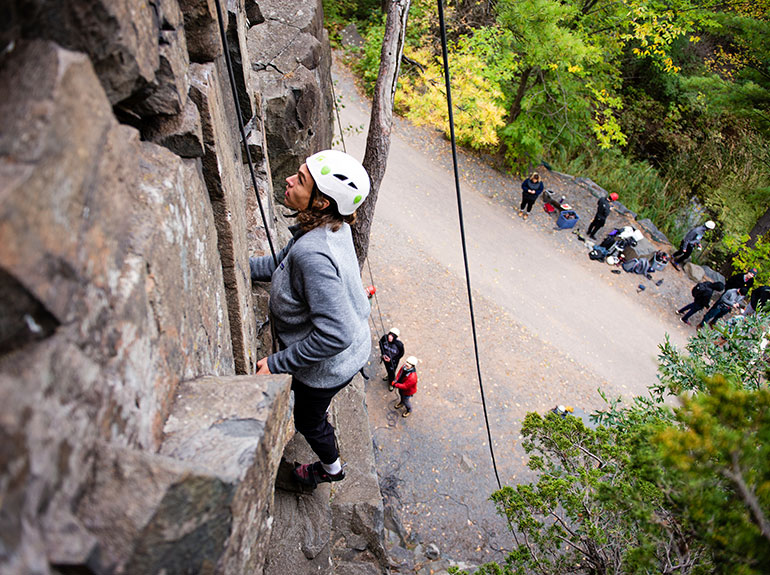 A student on belay rock climbs as students observe below