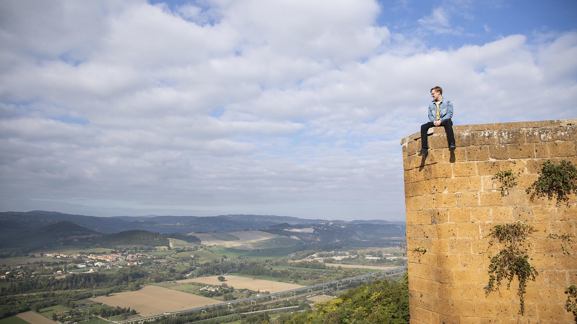A student sits on the edge of a tall tower overlooking a vast valley in Italy
