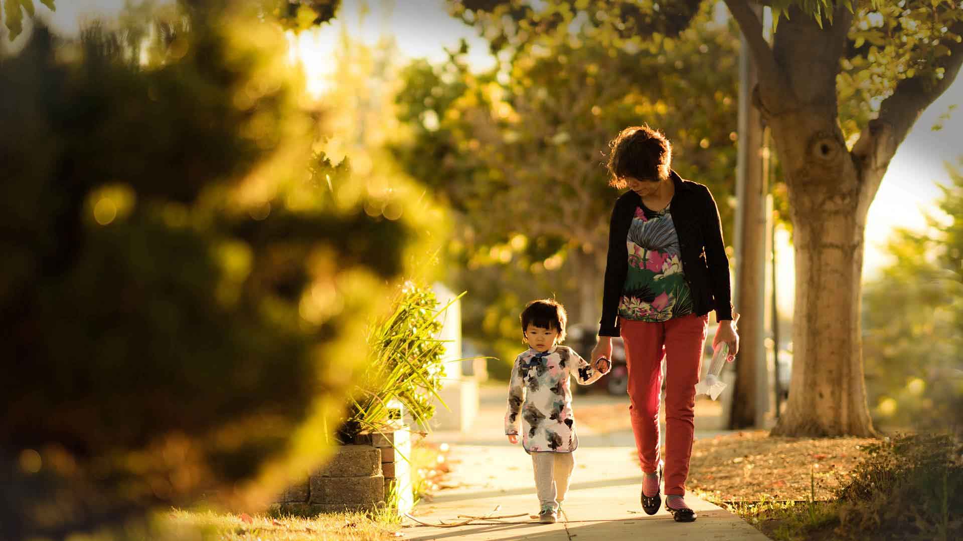 A woman holds a child's hand as they walk down the street at sunset.