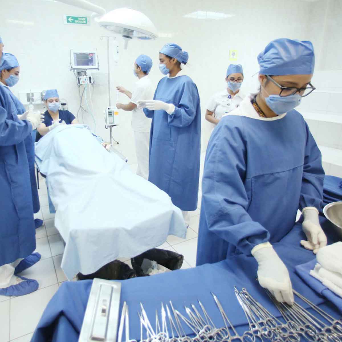 A physician assistant prepares surgical tools in a surgery room with a surgical team in the background. 
